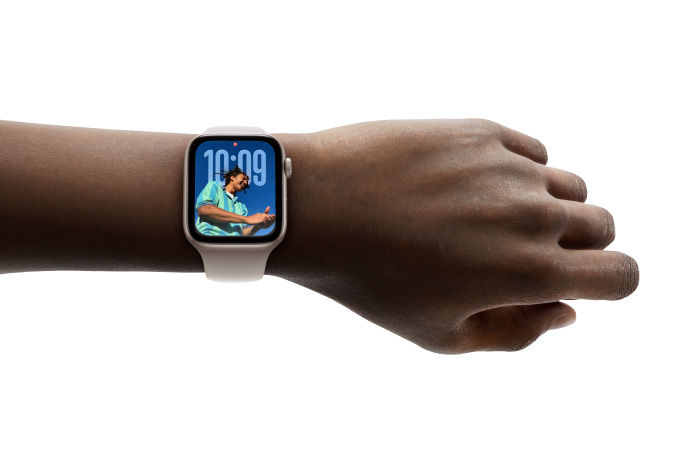Nærbillede af rosaguld Apple Watch med gummiurrem på et håndled. Skærmen viser Tid-appen i det nye Liquid Glass-design.