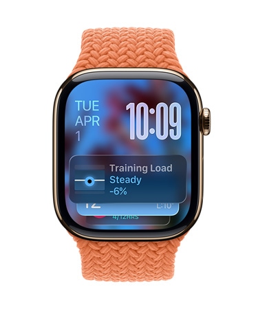 Rozéarany Apple Watch mandarinszínű fonott szíjjal. A számlap bal felső sarkában a dátum jelenik meg, a jobb felső sarokban az időt mutató számjegyek, alul pedig az Edzésterhelés widgetje. Mindegyik elem áttetsző Liquid Glass-megjelenést használ.