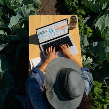 En kvinde med en bredskygget hat vist ovenfra. Hun sidder ved et bord i en have og arbejder på en virksomhedspræsentation på en MacBook.