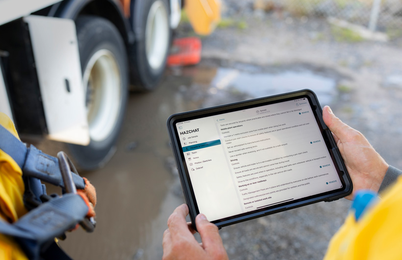 iPad tenu entre les mains d’une personne sur un lieu de travail près d’une flaque de boue. On voit à l’écran l’application HazChat affichant les catégories Job Details, Planning, Hazards and Controls